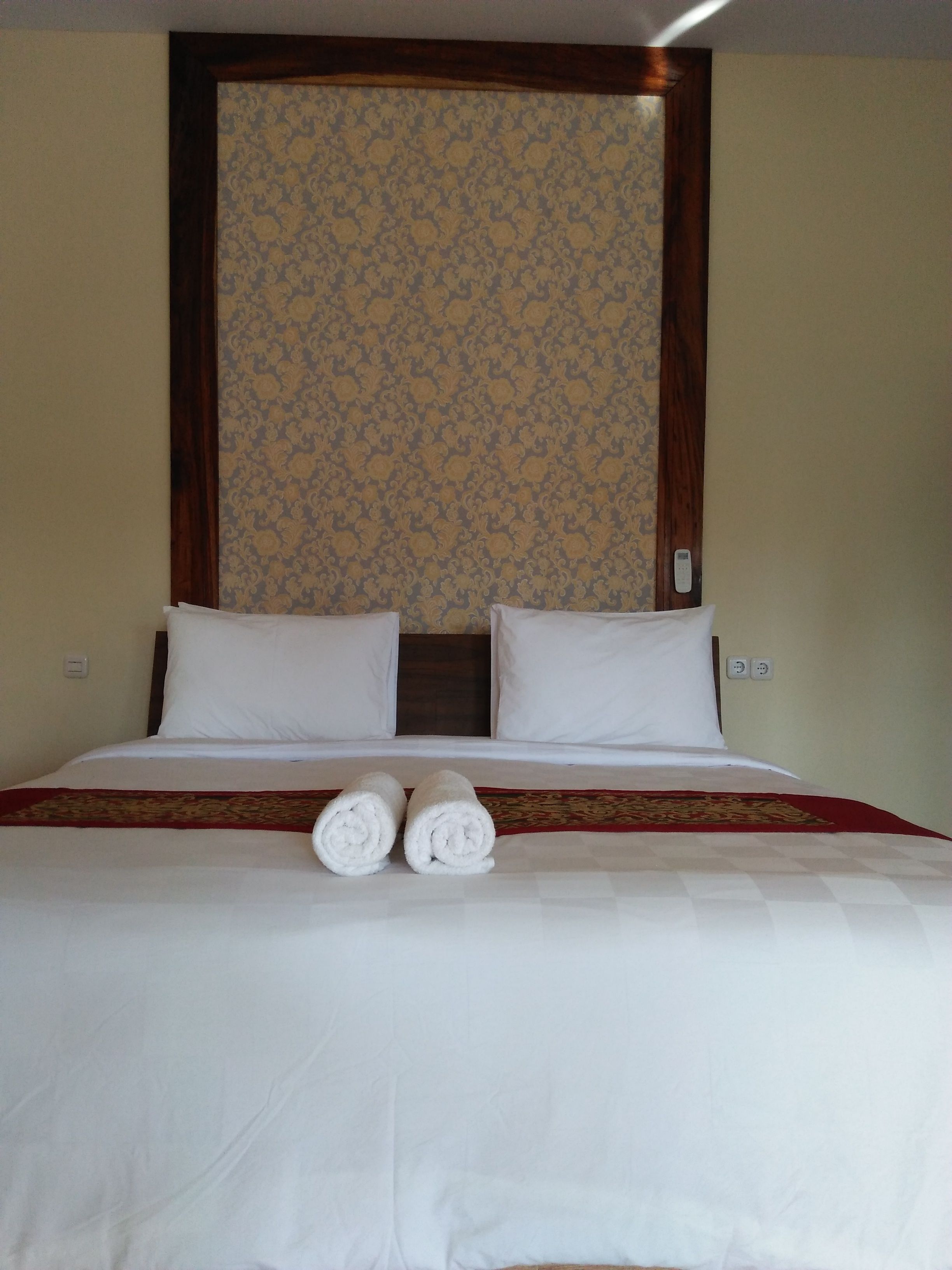 A neatly made bed with two rolled towels, a patterned wall, and a wooden headboard.