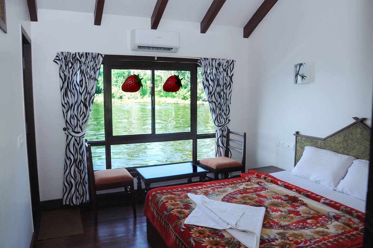 Un dormitorio con cama, una mesa y una silla. Hay una ventana con cortinas y vistas al exterior con vegetación.