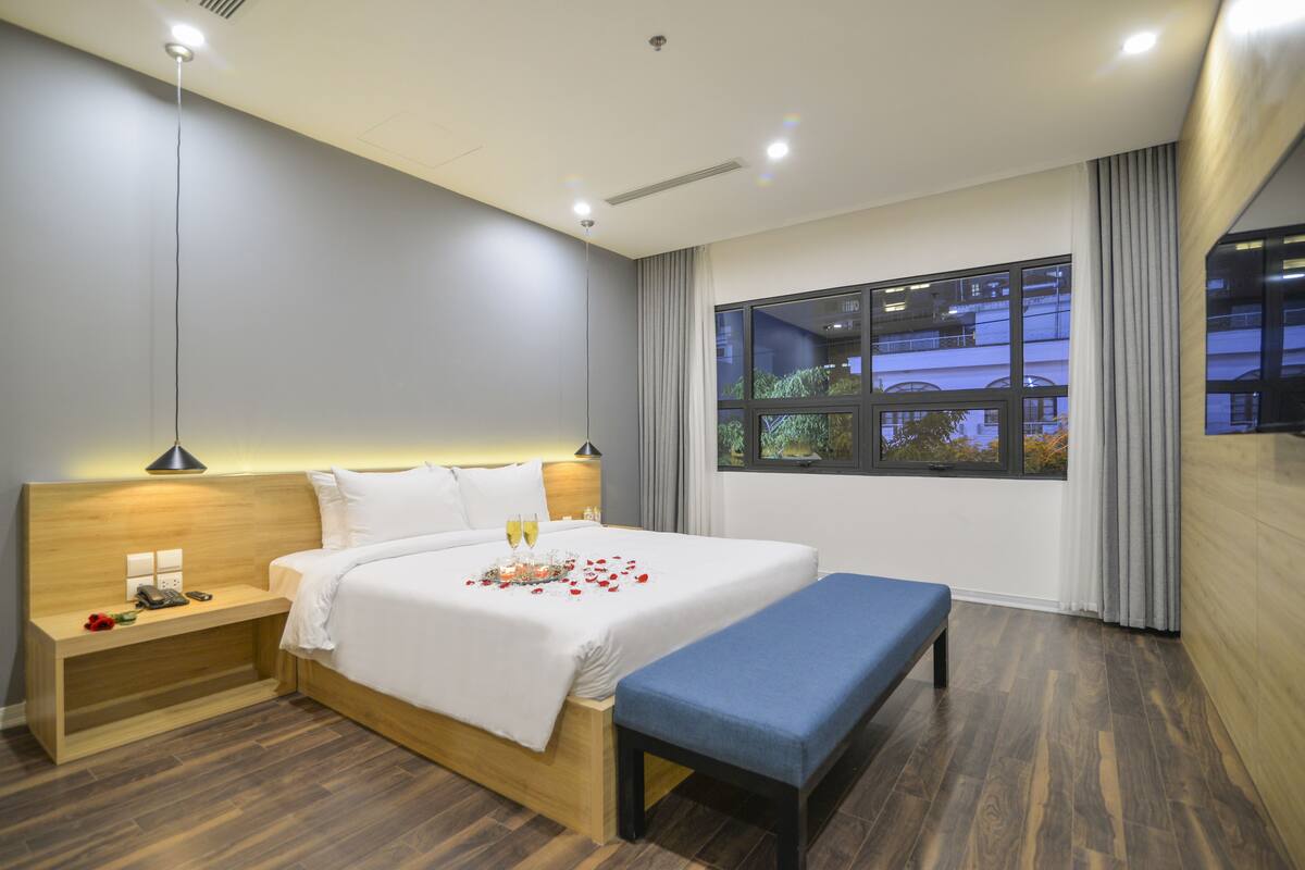 A modern hotel room with a wooden headboard, a large bed, a bedside table with a lamp, a bench, and a large window with a view of buildings.