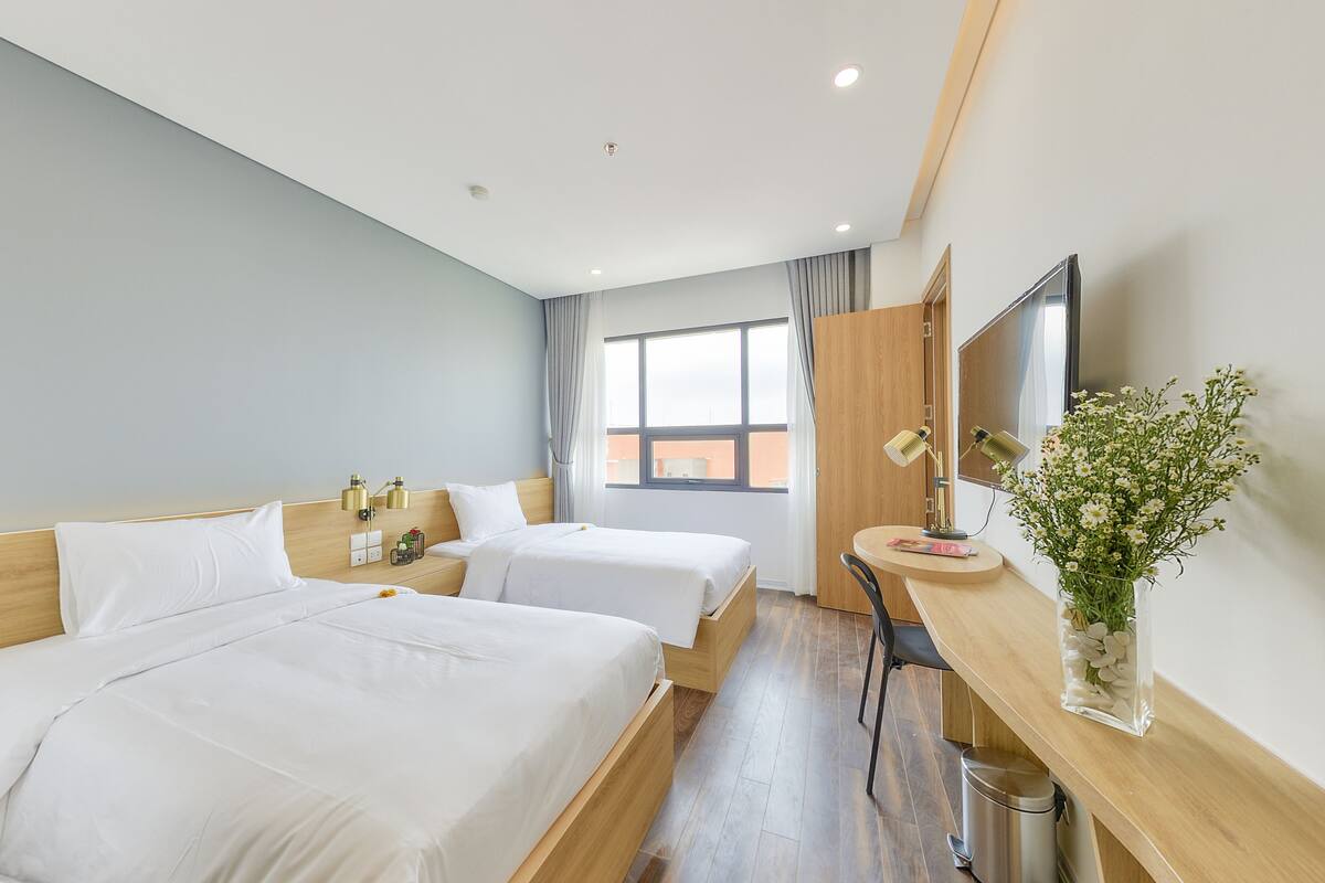 A hotel room with two beds, a desk with a lamp, a TV, and a window with curtains.