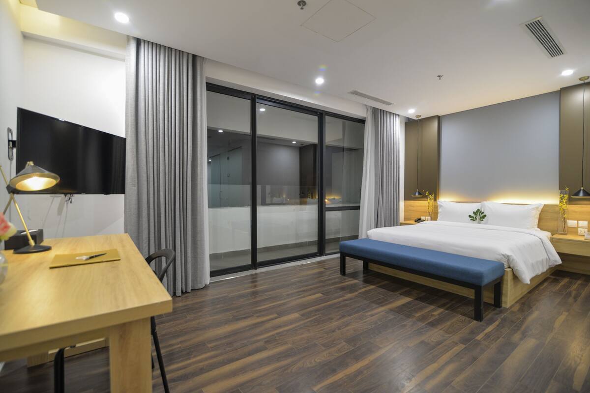 A hotel room with a wooden desk, a bed with a blue bench, a TV, and a sliding glass door leading to a balcony.
