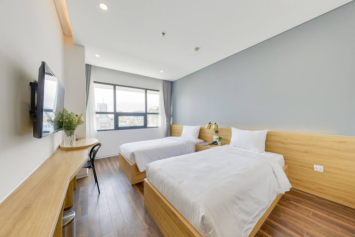 A modern hotel room with two beds, a desk, a TV, and a window with a city view.