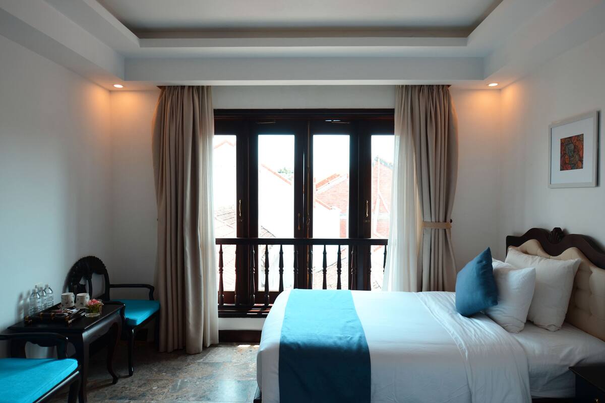 A hotel room with a bed, a desk, and a view through a window with curtains.