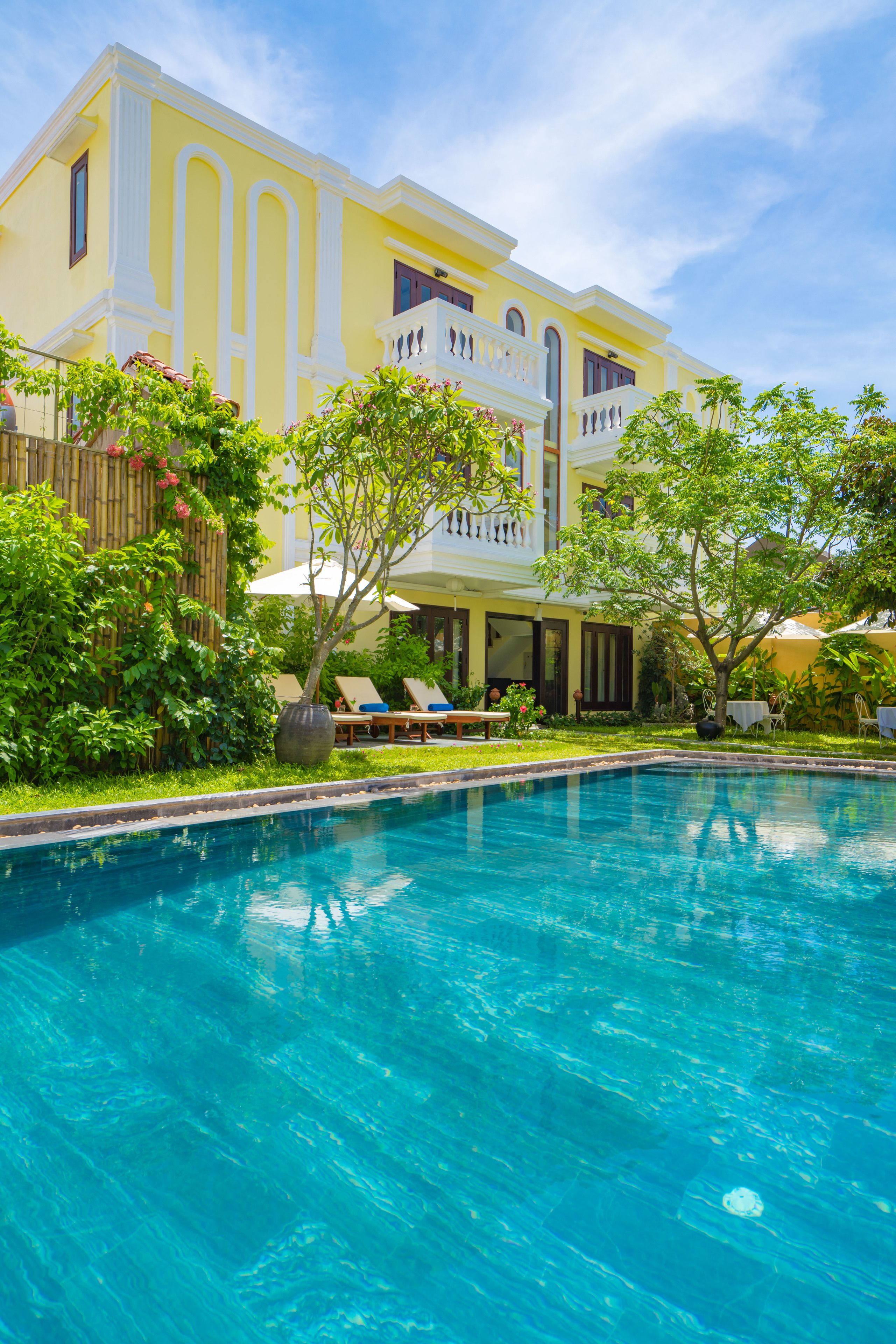 A large yellow building with a swimming pool, surrounded by lush greenery and outdoor seating areas.