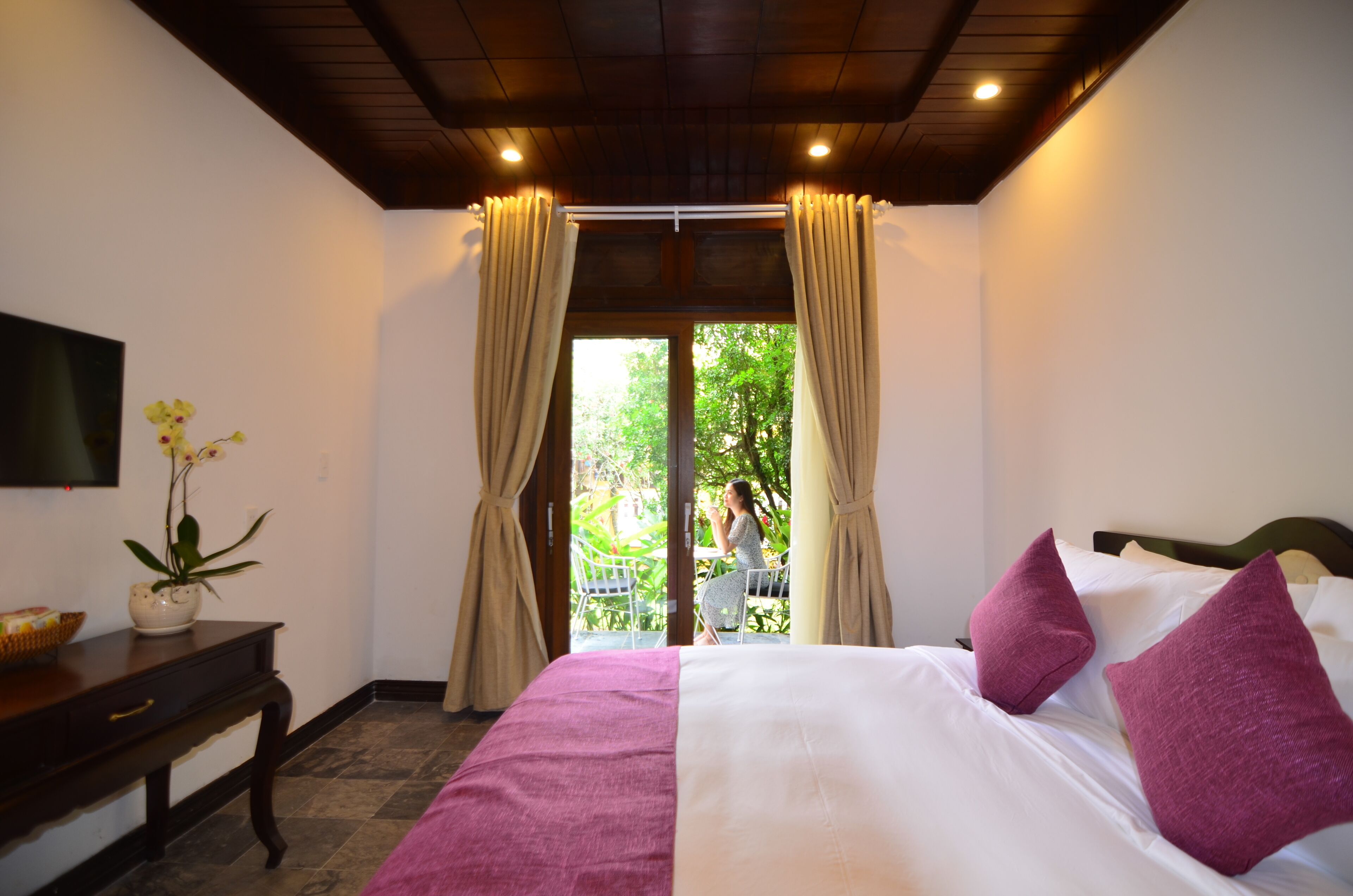 Ein Hotelzimmer mit einem Bett, einem an der Wand montierten Fernseher, einem kleinen Tisch mit Blumenvase und einem Fenster mit Vorhängen.