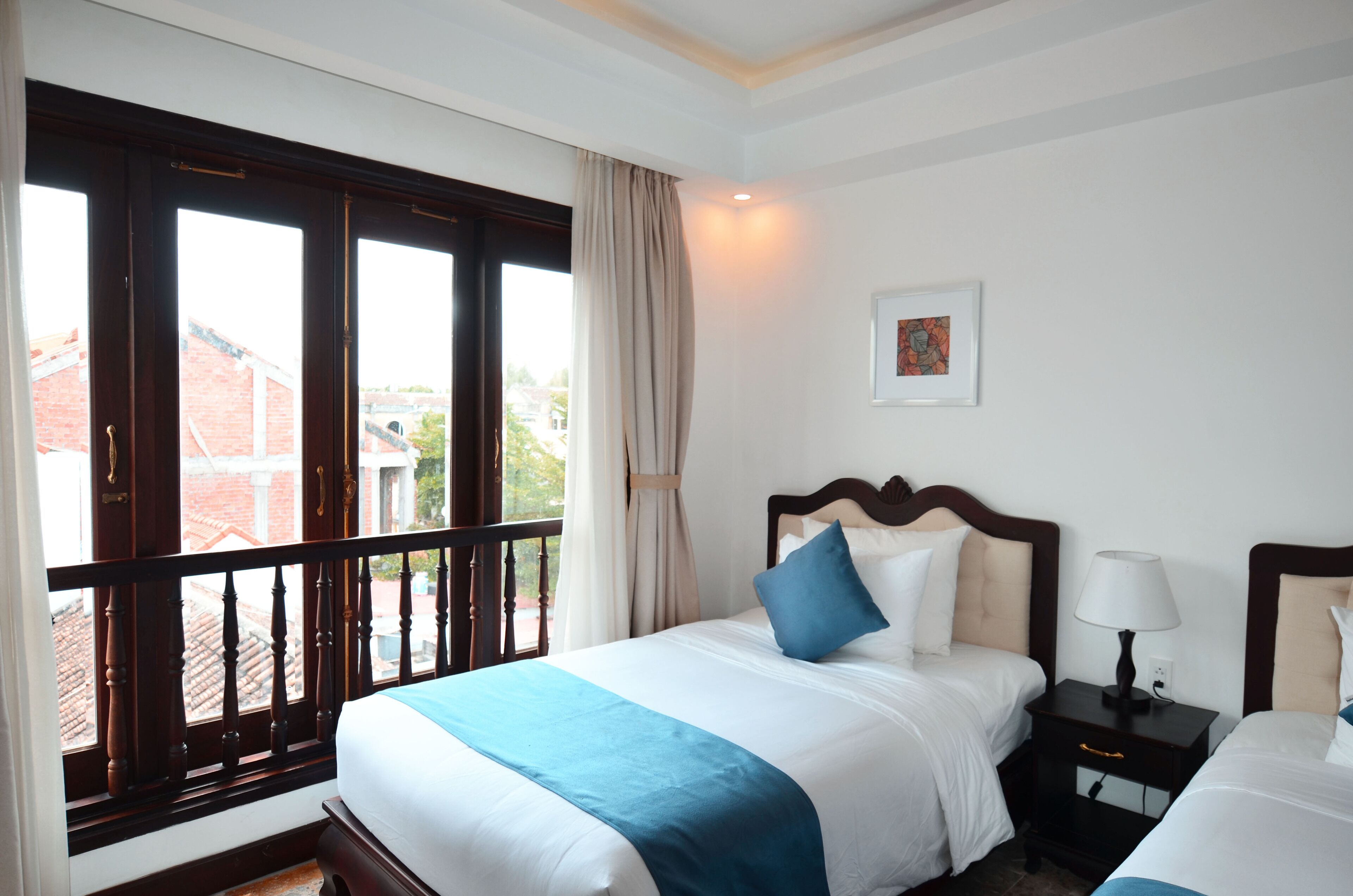 A hotel room with a large window, a bed with a blue accent, a bedside table with a lamp, and a framed picture on the wall.