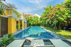 A swimming pool with a tiled deck, surrounded by lush greenery and a yellow building with a tiled roof. - Hoian Central Hotel (Da Nang)