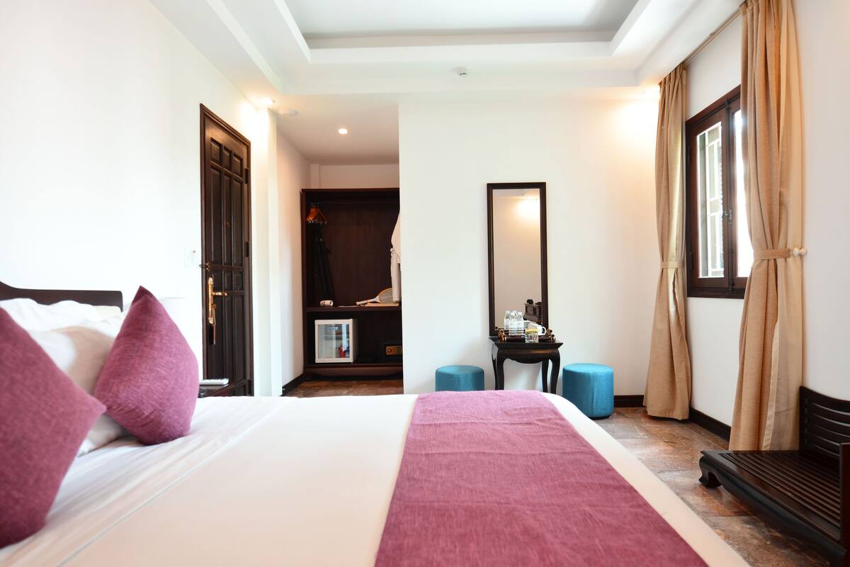 A hotel room with a large bed, a wardrobe, a mirror, a small table, and a window with curtains.