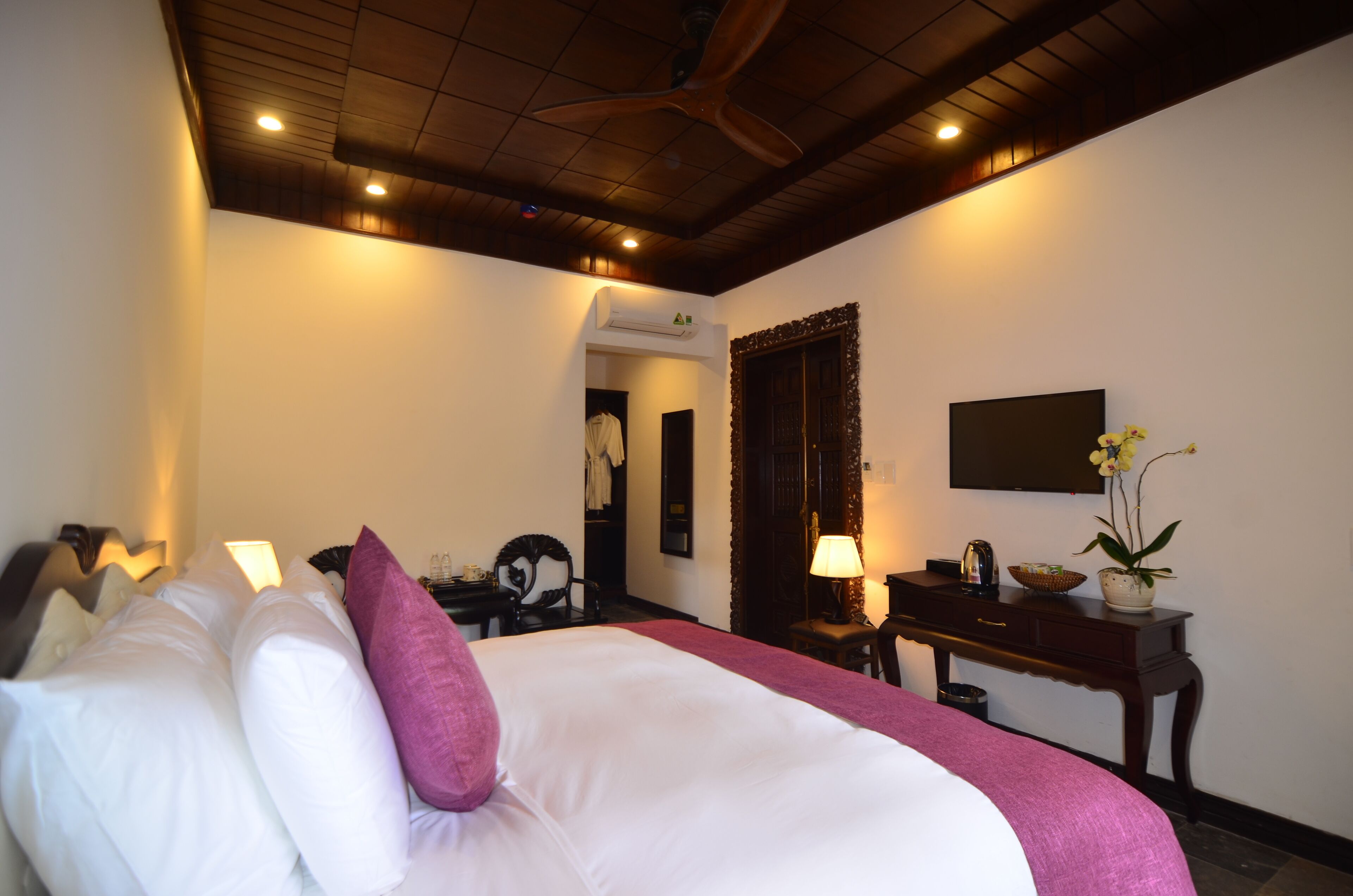 A hotel room with a large bed, a desk, a chair, a TV, and a ceiling fan.
