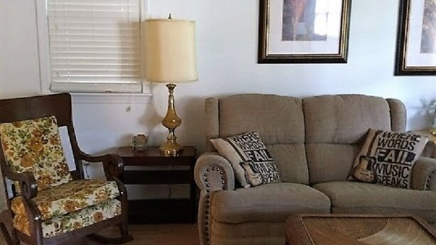 Un salon comprenant un canapé, un fauteuil et une table basse. Deux tableaux encadrés ornent le mur, une lampe se trouve sur une table d’appoint, et une fenêtre est équipée de stores.