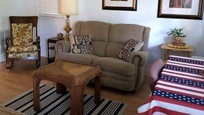 Un salon comprenant un canapé, un fauteuil, une table basse et une petite table avec une lampe. Des tableaux encadrés ornent le mur, et il y a une fenêtre avec des stores.