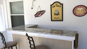 A bar area with a granite countertop, two bar stools, and wall decorations including a surfboard and a clock sign. - 1 Block to the Beach, Fully Fenced in Backyard, Golf Cart Included (Myrtle Beach)