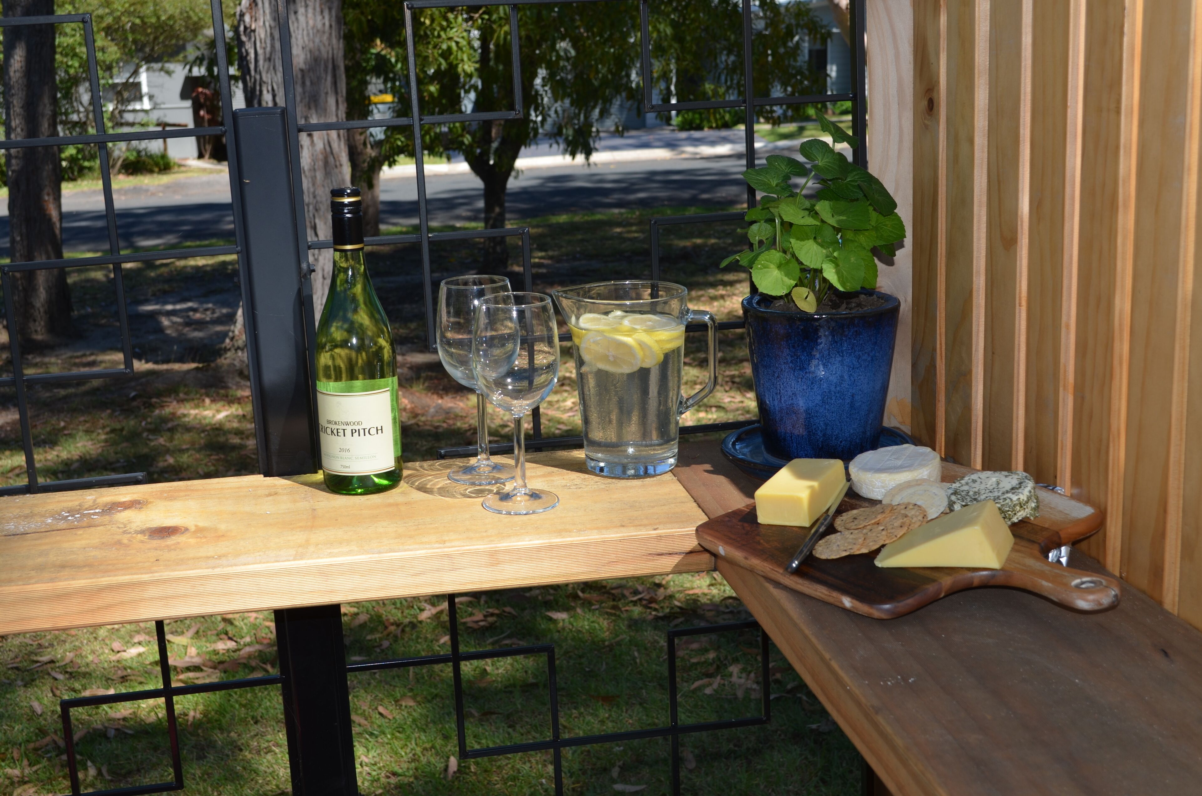Una mesa con una botella de vino, copas, una jarrón con rodajas de limón, una planta en maceta y una tabla de madera con queso y galletas.