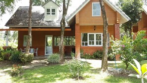 Een tweelaags huis met een veranda, een kleine garage en een verzorgde tuin met diverse planten en gazon.