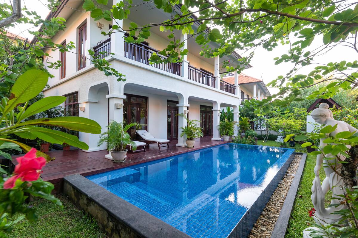 A two-story house with a swimming pool, surrounded by lush greenery and potted plants.