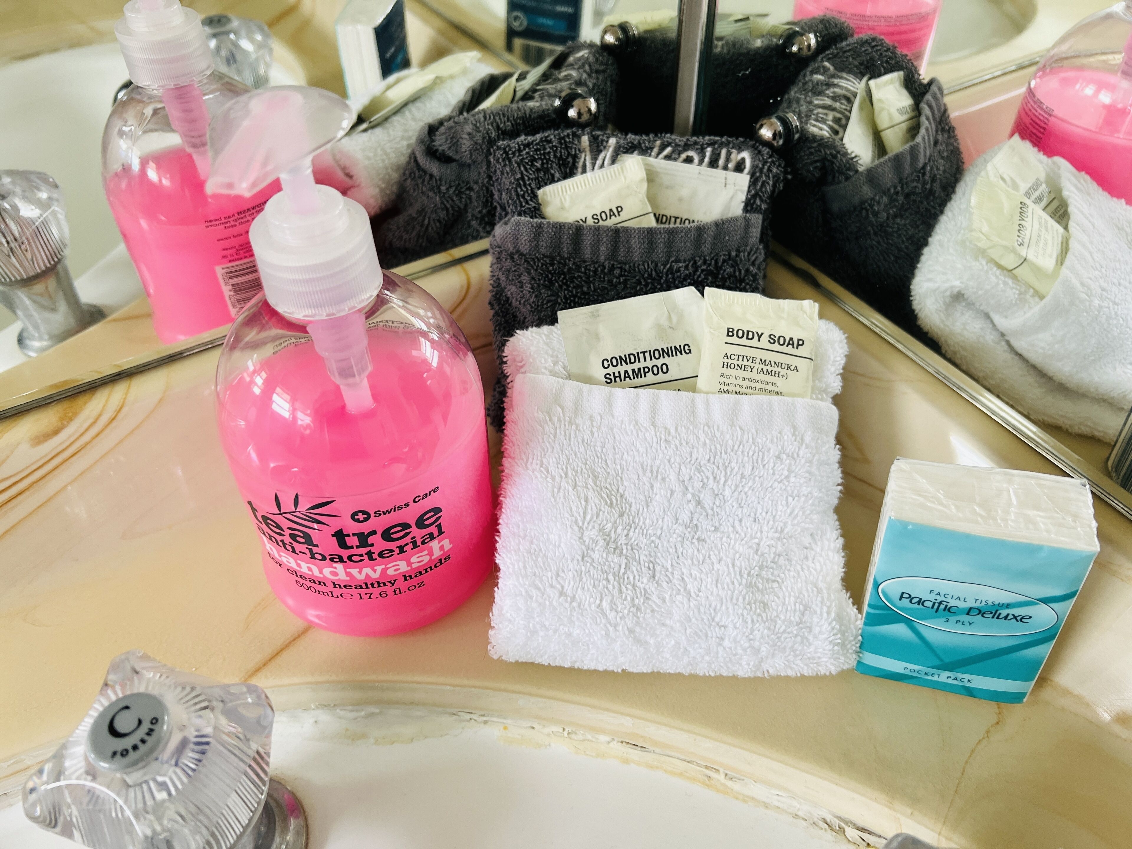 A bathroom vanity with a white towel, a pink bottle of body soap, a black towel, and a facial tissue pack.
