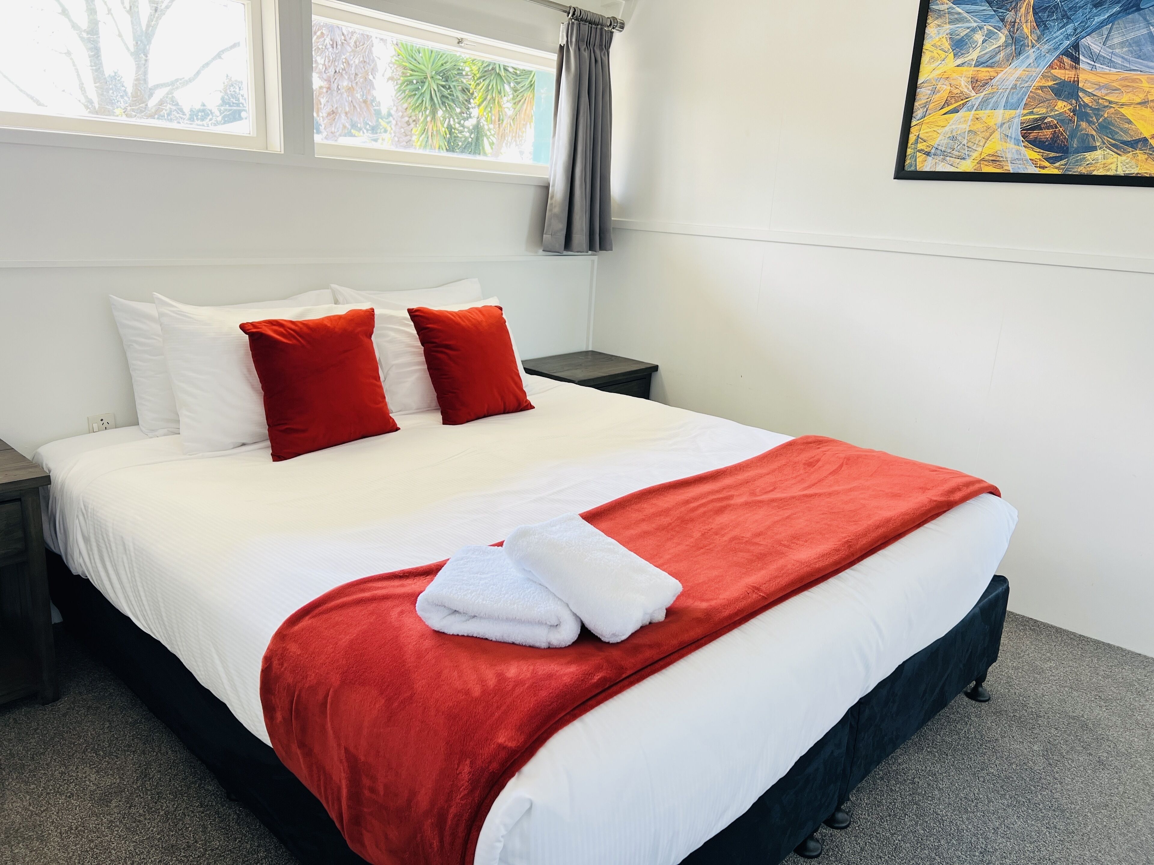 Een net opgemaakt bed met witte en rode beddengoed, een grijze nachtkastje en een raam met gordijnen.