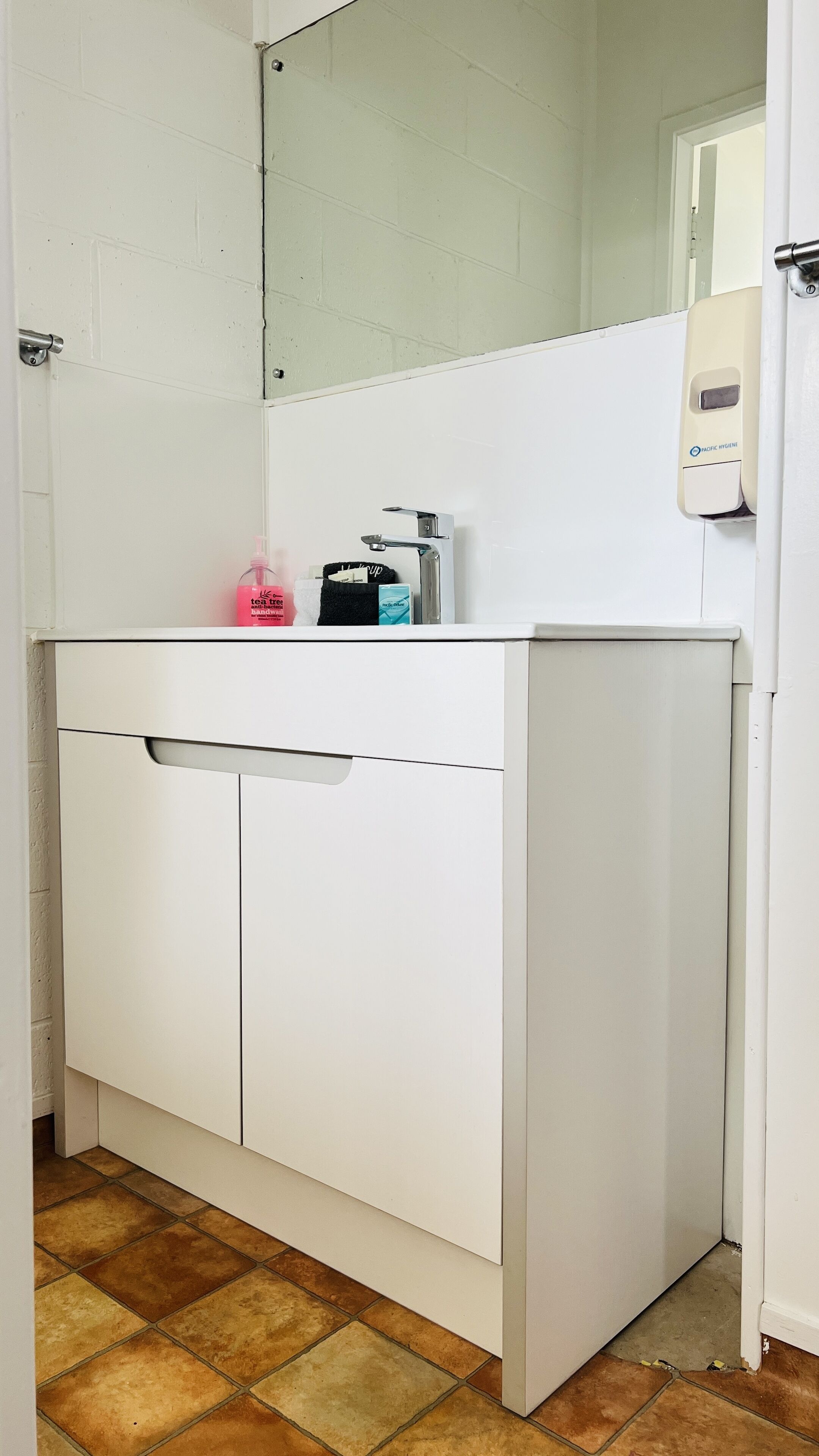 A white bathroom with a sink, mirror, and soap dispenser.
