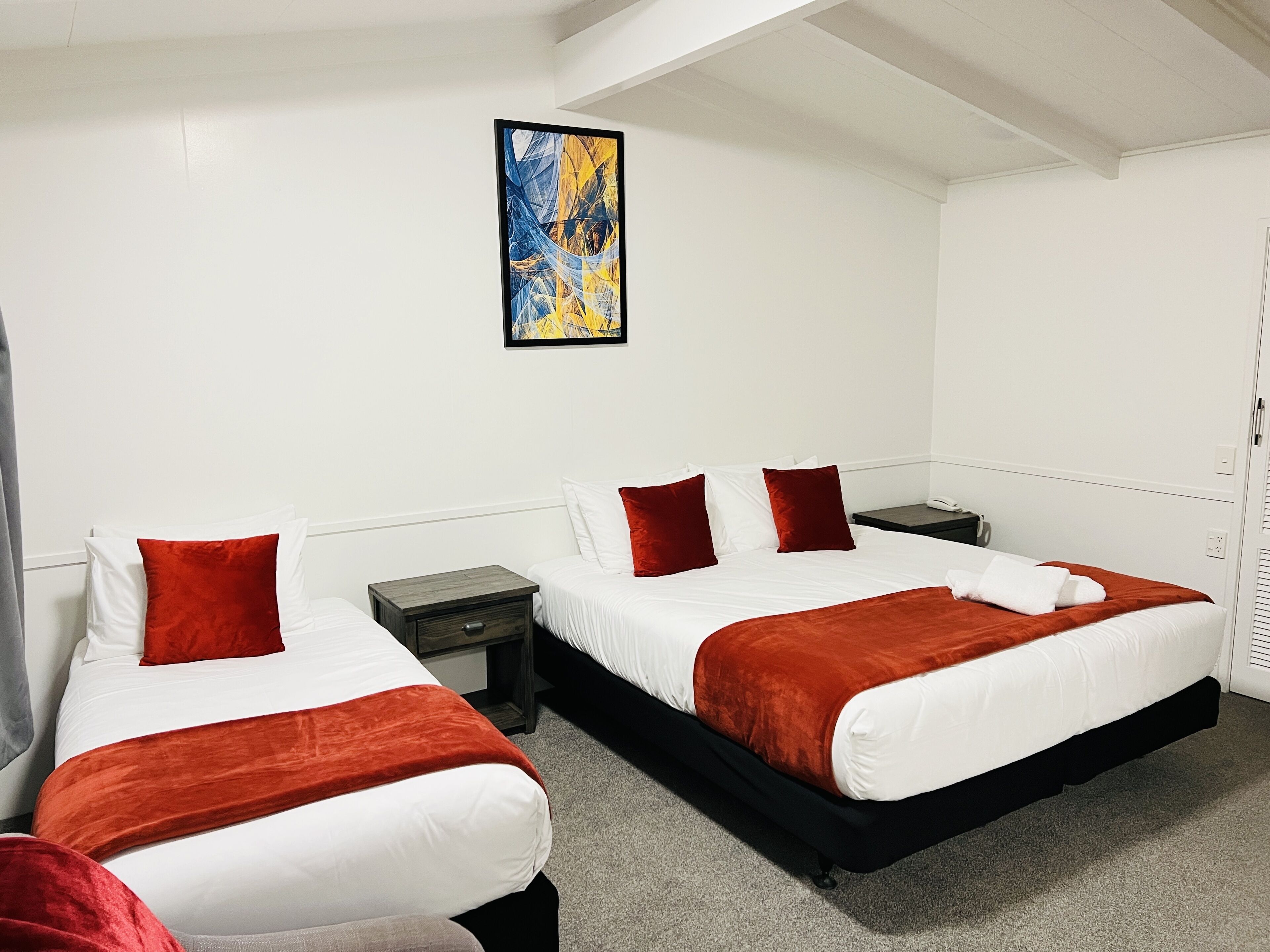 Habitación de hotel con dos camas, cada una con ropa blanca y almohadas rojas, una mesita de noche pequeña, un buró, un cuadro en la pared y una ventana con persianas.