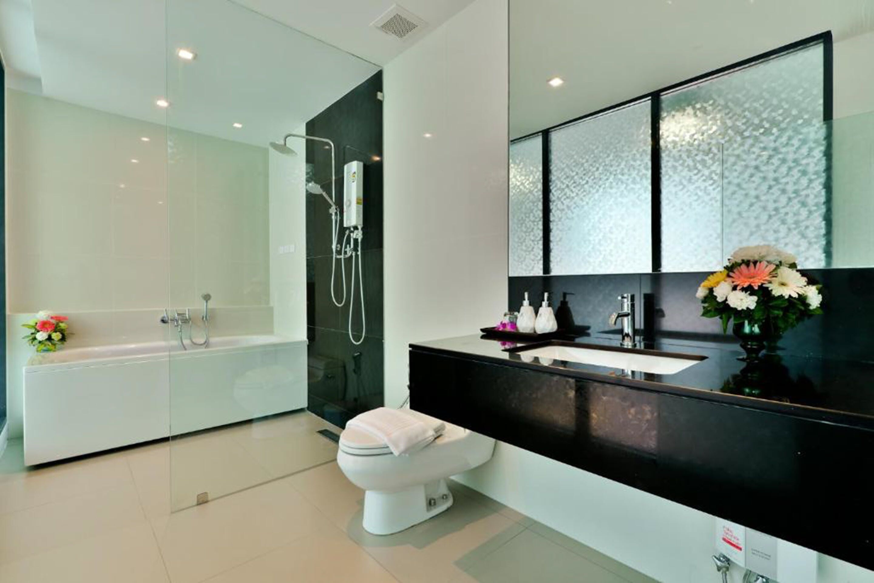 Un baño moderno con ducha de vidrio, inodoro blanco, tocador negro con lavamanos y un espejo grande.