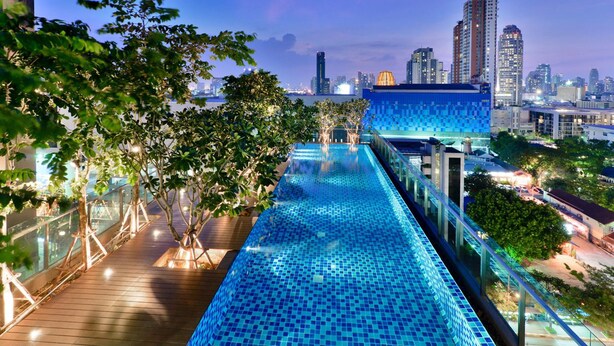 A rooftop infinity pool with a city skyline view, surrounded by trees and modern buildings.