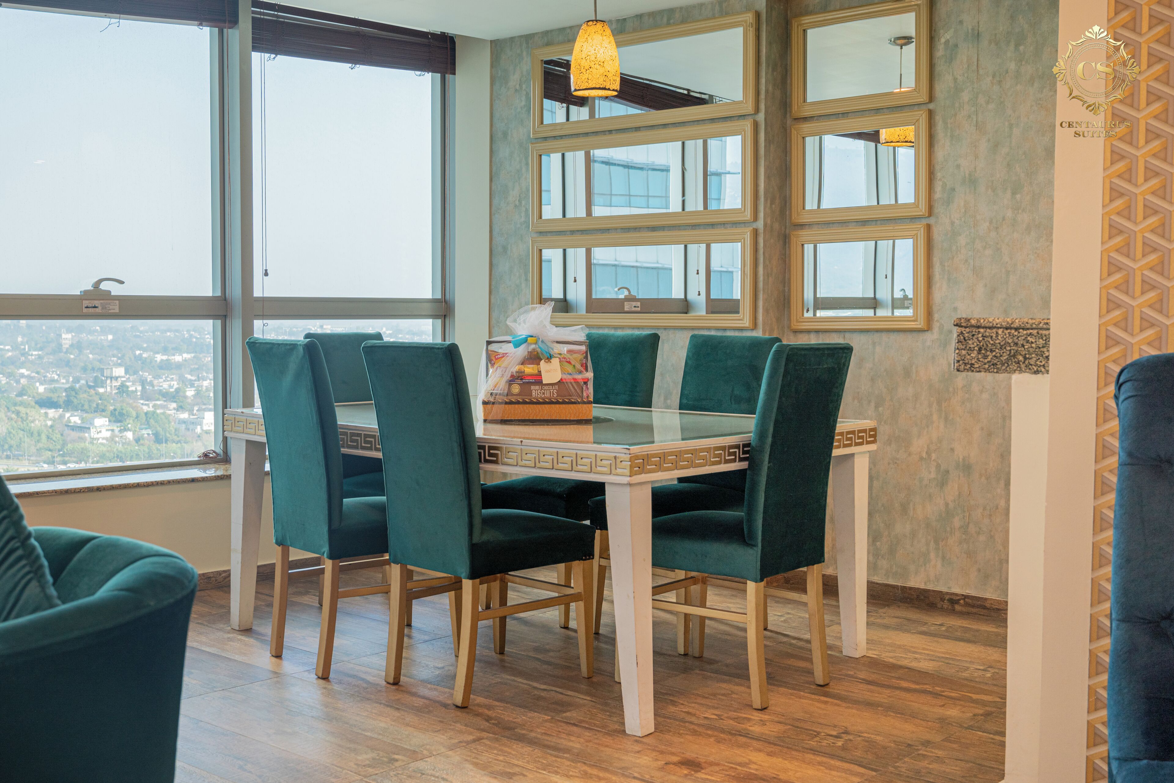 A dining area with a glass table, green upholstered chairs, and a view through large windows.