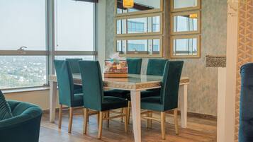 A dining area with a glass table, green upholstered chairs, and a view through large windows.