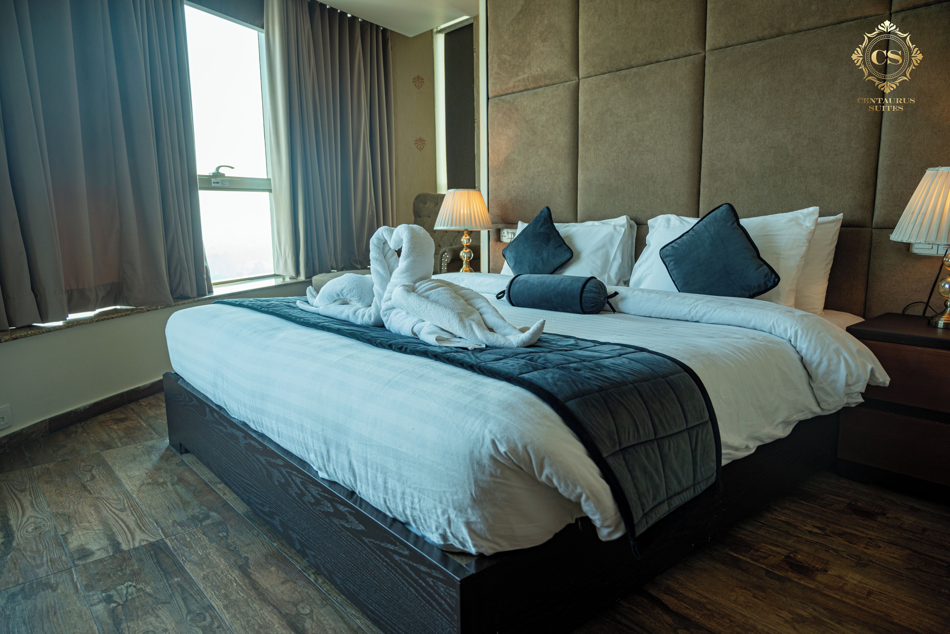 A hotel room with a large bed, a headboard, a bedside table, a lamp, and a window with curtains.