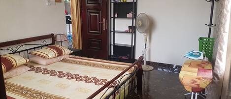 Un dormitorio con una cama, un ventilador de pie, un cesto de ropa verde y una puerta que da a otra habitación.
