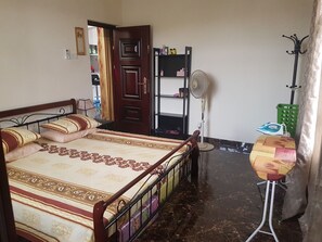 Une chambre à coucher avec un lit, un ventilateur sur pied, un panier à linge vert et une porte donnant sur une autre pièce.