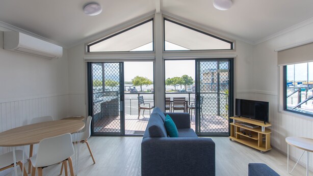 A modern living room with a large window, a sofa, a TV stand, and a dining area. - Jacobs Well Tourist Park (Jacobs Well)