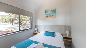 A hotel room with a large bed, a window with a view of the outside, a painting on the wall, and two bedside tables with lamps. - Jacobs Well Tourist Park (Jacobs Well)