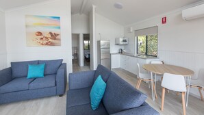 A modern living room with a grey sofa and teal cushions, a wooden dining table with white chairs, and a kitchen area with white cabinets and a microwave. - Jacobs Well Tourist Park (Jacobs Well)