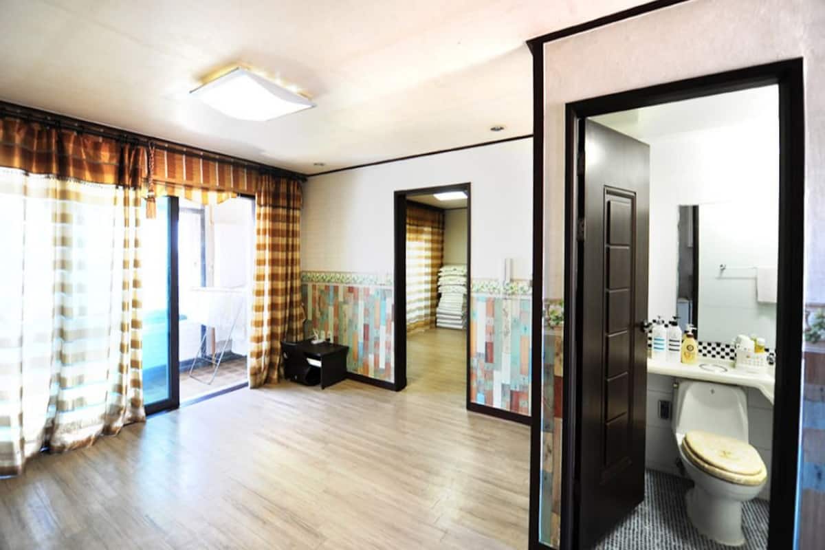 A hotel room with a wooden floor, a black chair, a small table, and a bathroom with a toilet and sink.
