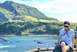A person sitting on a wooden ledge overlooking a coastal area with green hills and a clear blue sea.