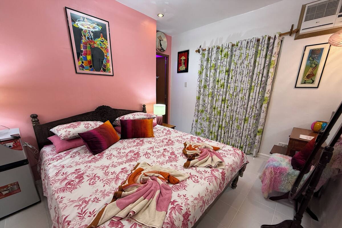 A bedroom with a bed, pillows, a bedside lamp, a mirror, and a curtain.