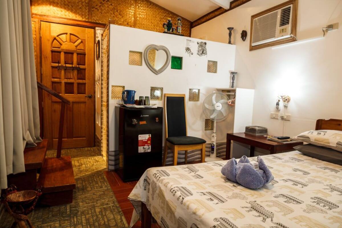 A hotel room with a bed, a desk, a chair, a small table, a fan, and a wooden door.