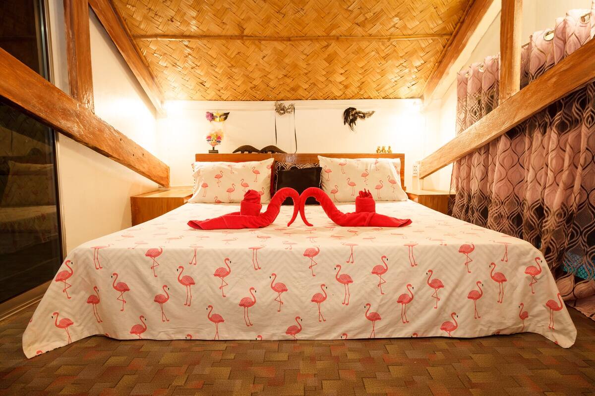 A hotel room with a bed featuring a flamingo pattern bedspread, wooden headboard, and a woven ceiling.
