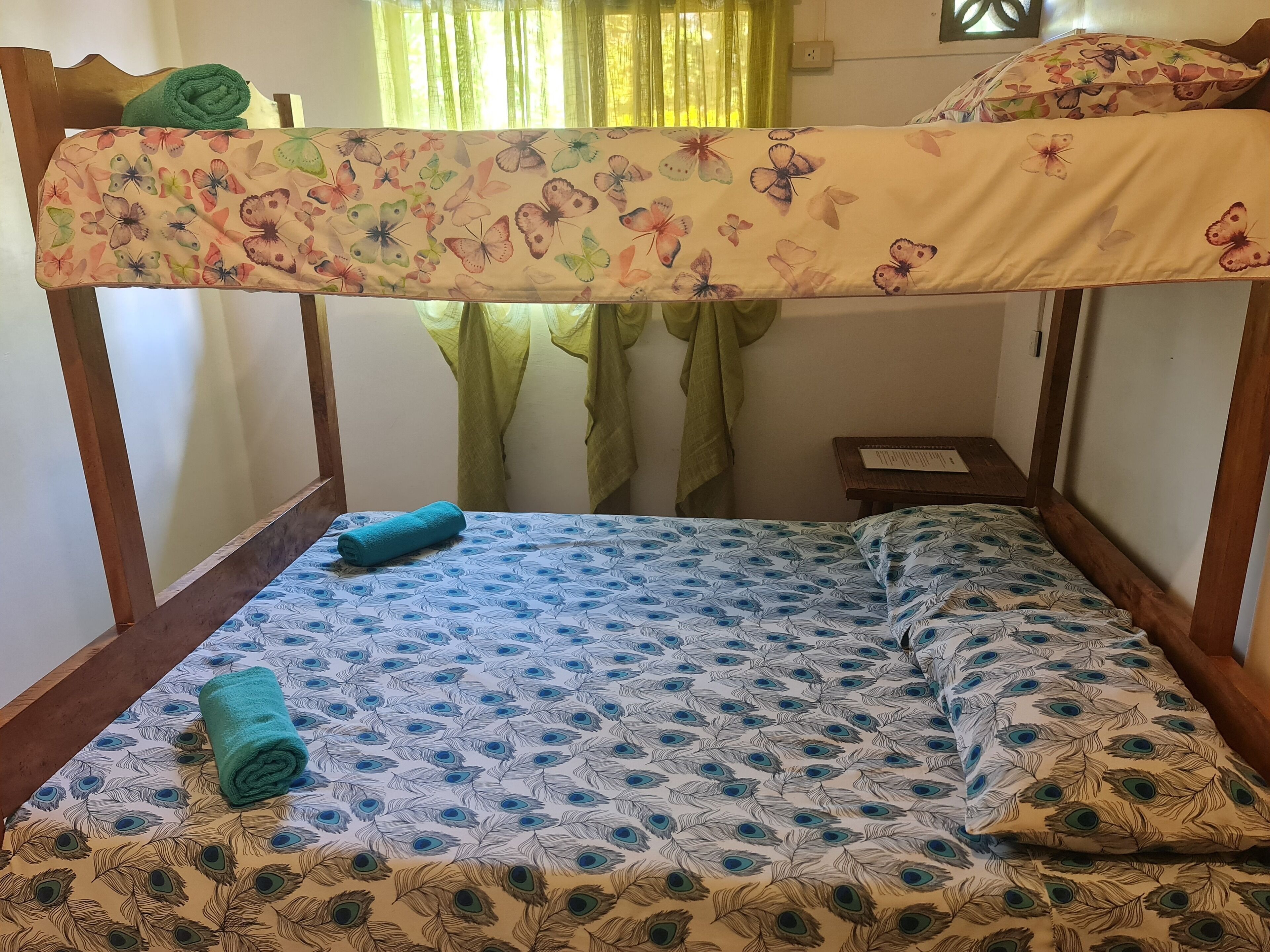 A single bed with a peacock feather pattern, a green towel, and a floral blanket.