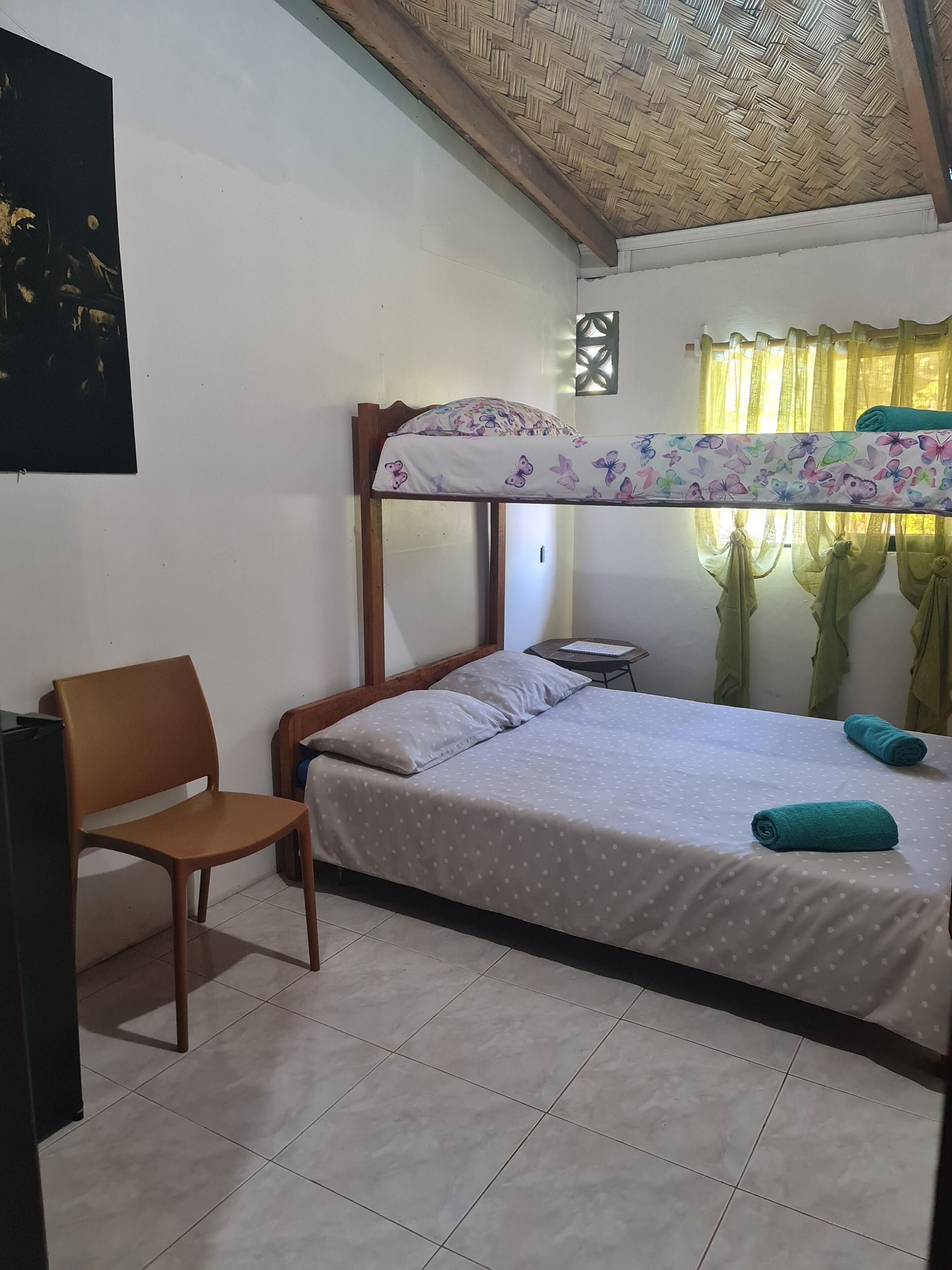 A bunk bed with a floral-patterned top sheet, a single bed with a polka-dotted sheet, a wooden chair, and a small table with a lamp.