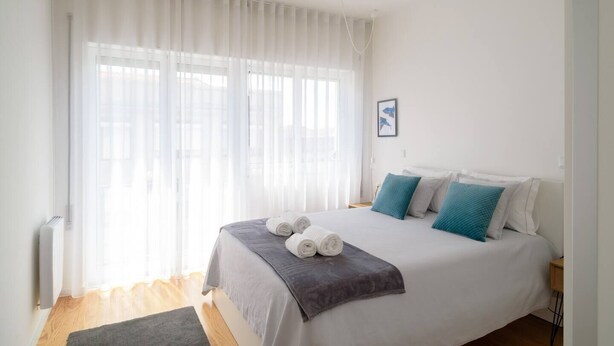 A neatly made bed with rolled towels, a grey blanket, and teal pillows in a bright room with sheer curtains and a framed picture on the wall.