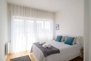 Una cama bien tendida con toallas enrolladas, una manta gris y almohadas turquesa en una habitación luminosa con cortinas de encaje y un cuadro en la pared.