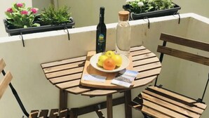 A small wooden table with a bottle, a plate with fruit, and a notebook on a balcony with potted plants.