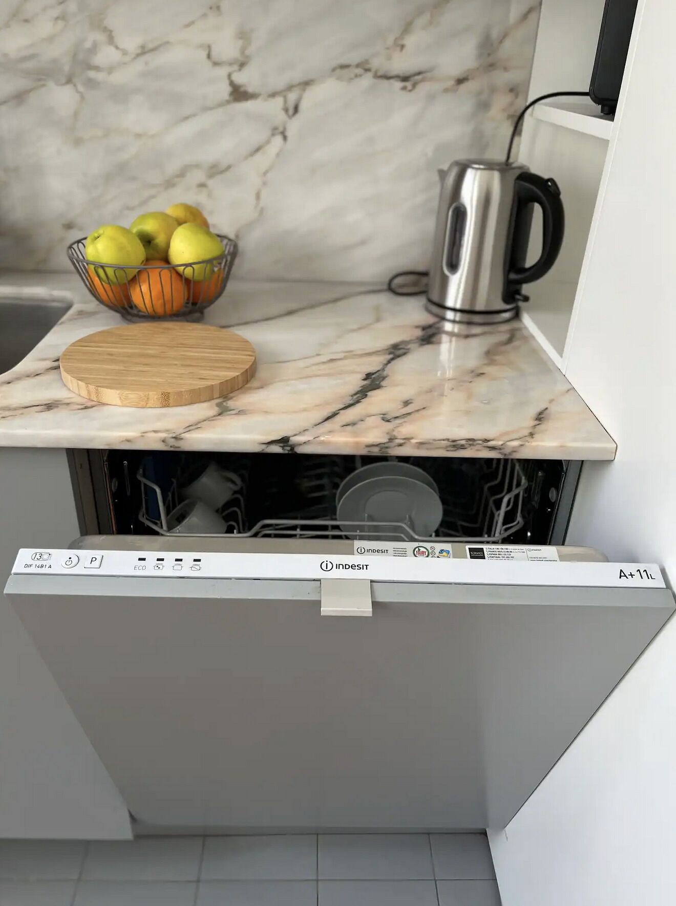 A kitchen with a marble countertop, a fruit bowl, a kettle, and a dishwasher.