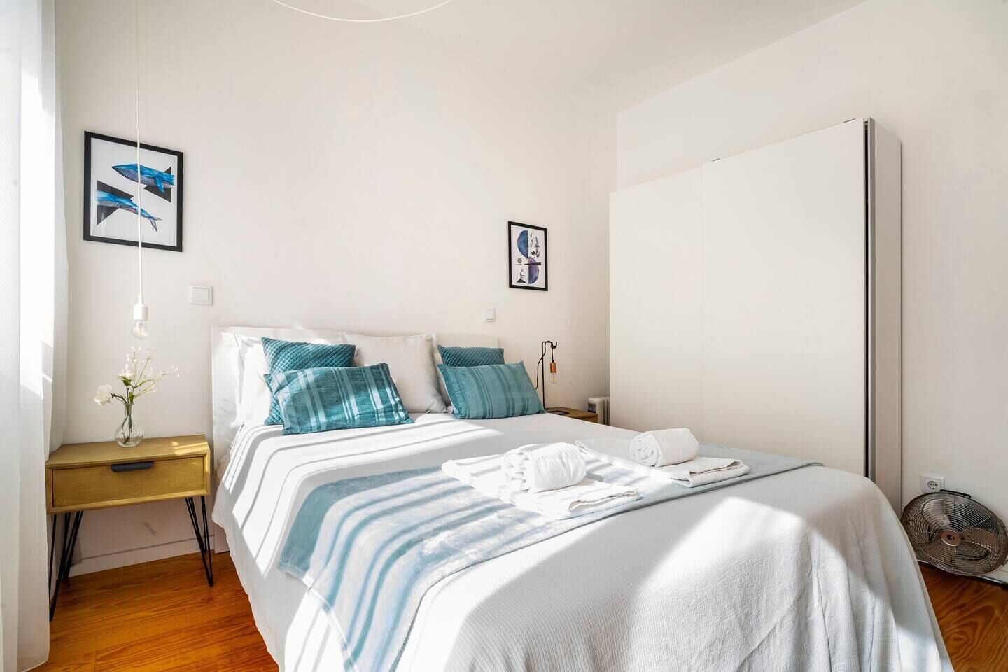 Une chambre à coucher comprenant un lit, des tables de chevet, une commode, un ventilateur fixé au mur et des œuvres d’art accrochées aux murs.