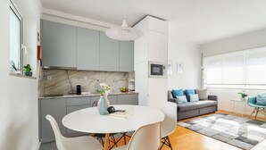 A modern kitchen with grey cabinets, a white dining table with four chairs, a white pendant light, and a grey and white checkered rug.