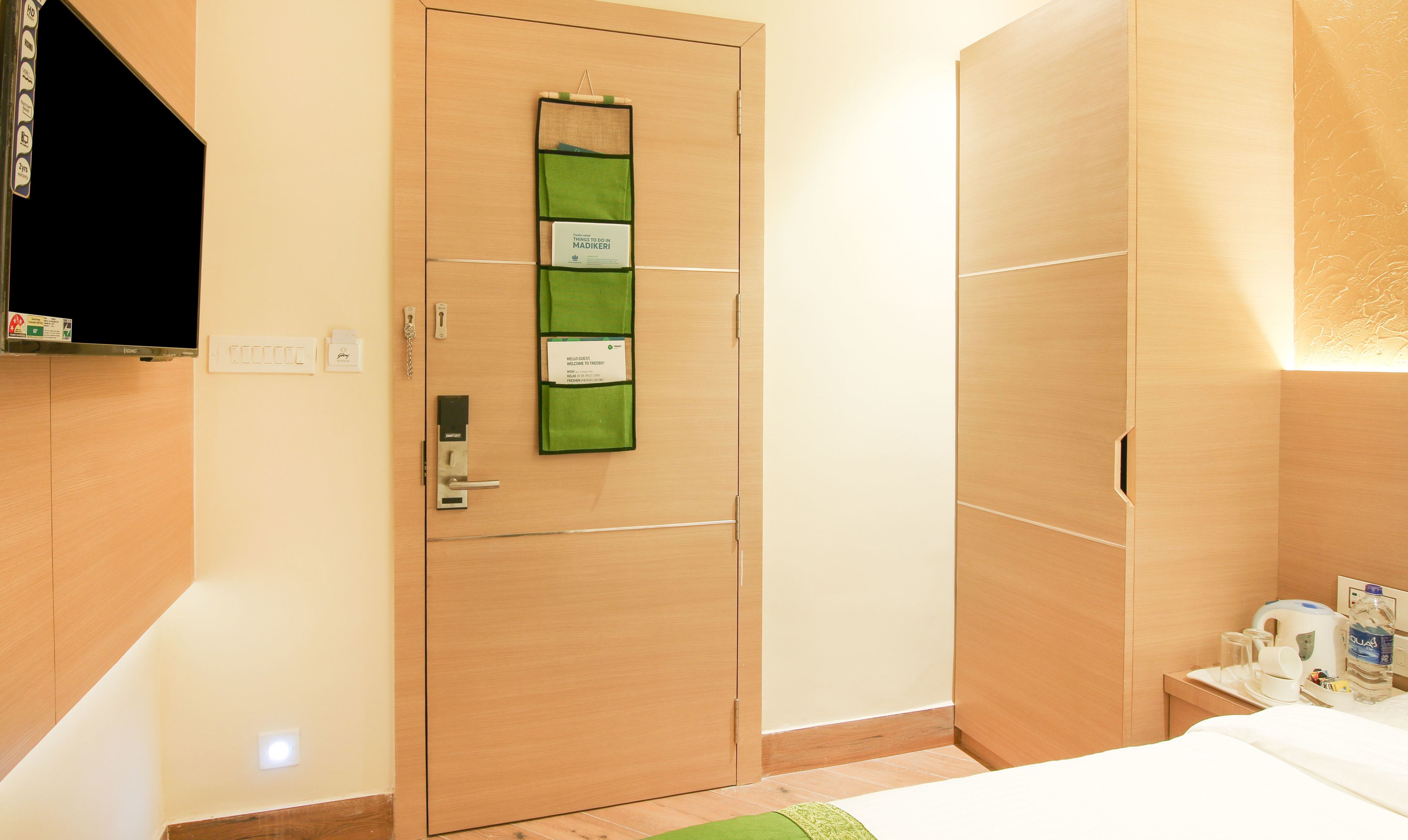 A hotel room with a wooden wardrobe, a green vertical sign on the door, a bed, a bedside table with a kettle, and a water bottle.