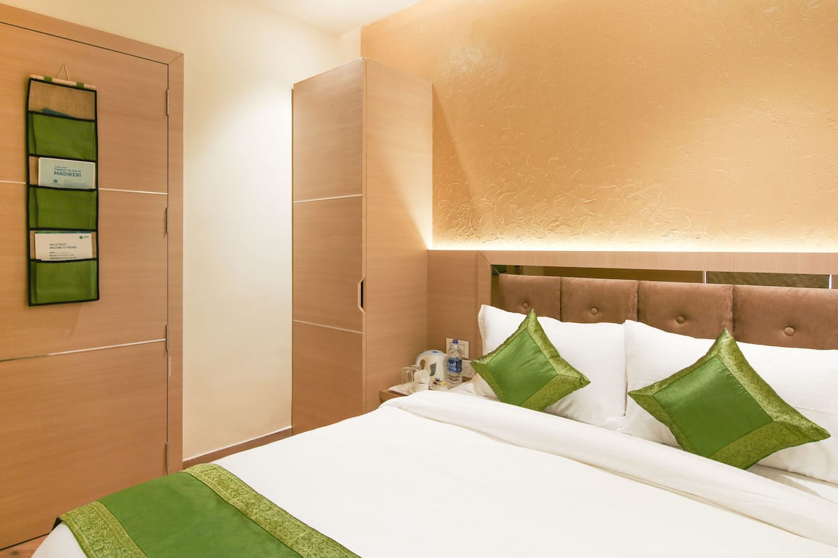 Habitación de hotel con un armario de madera, una cama con ropa blanca y almohadas verdes, y una lámpara de pared.