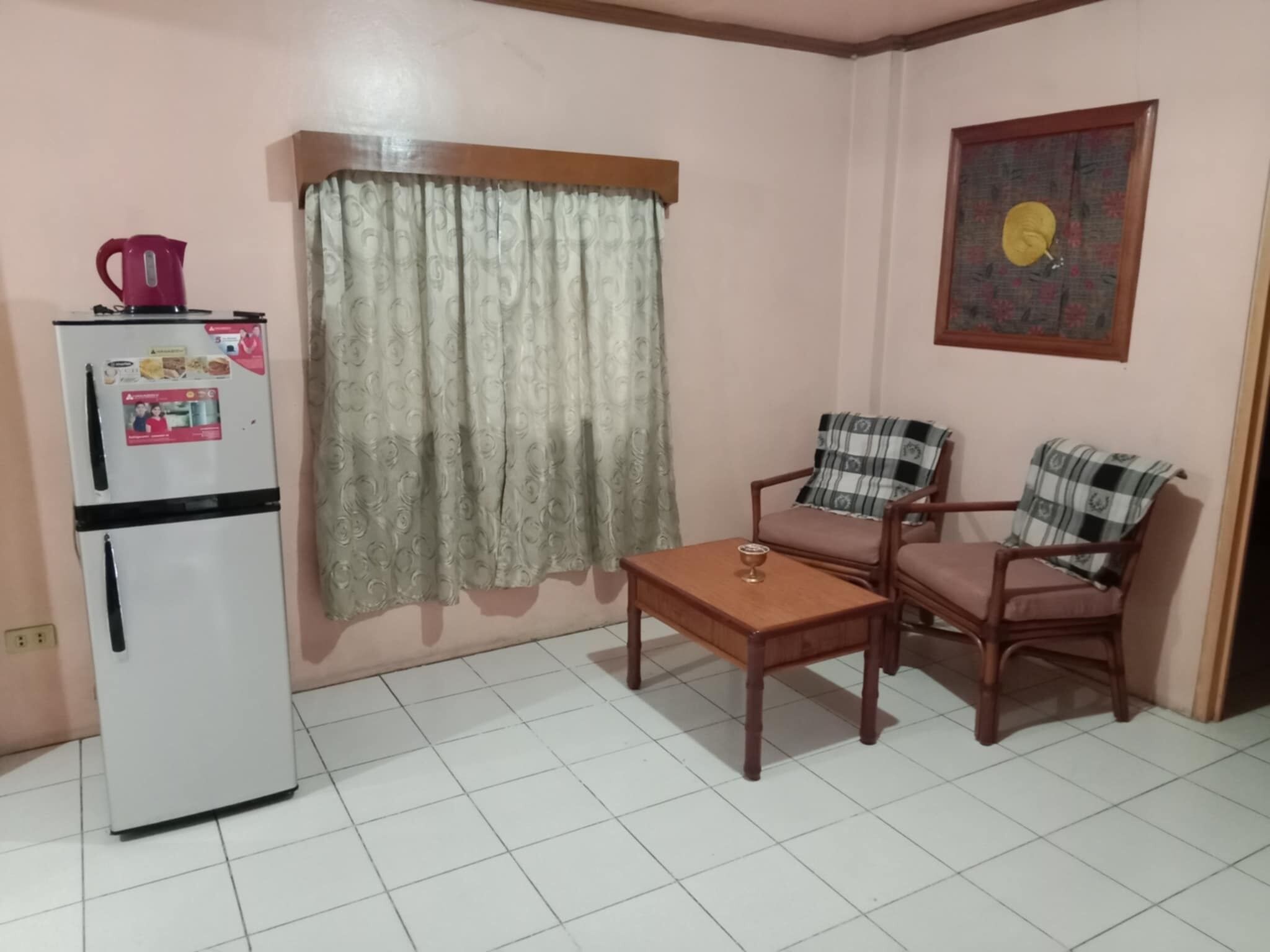 A small living area with a refrigerator, two chairs, a table, and a window with curtains.