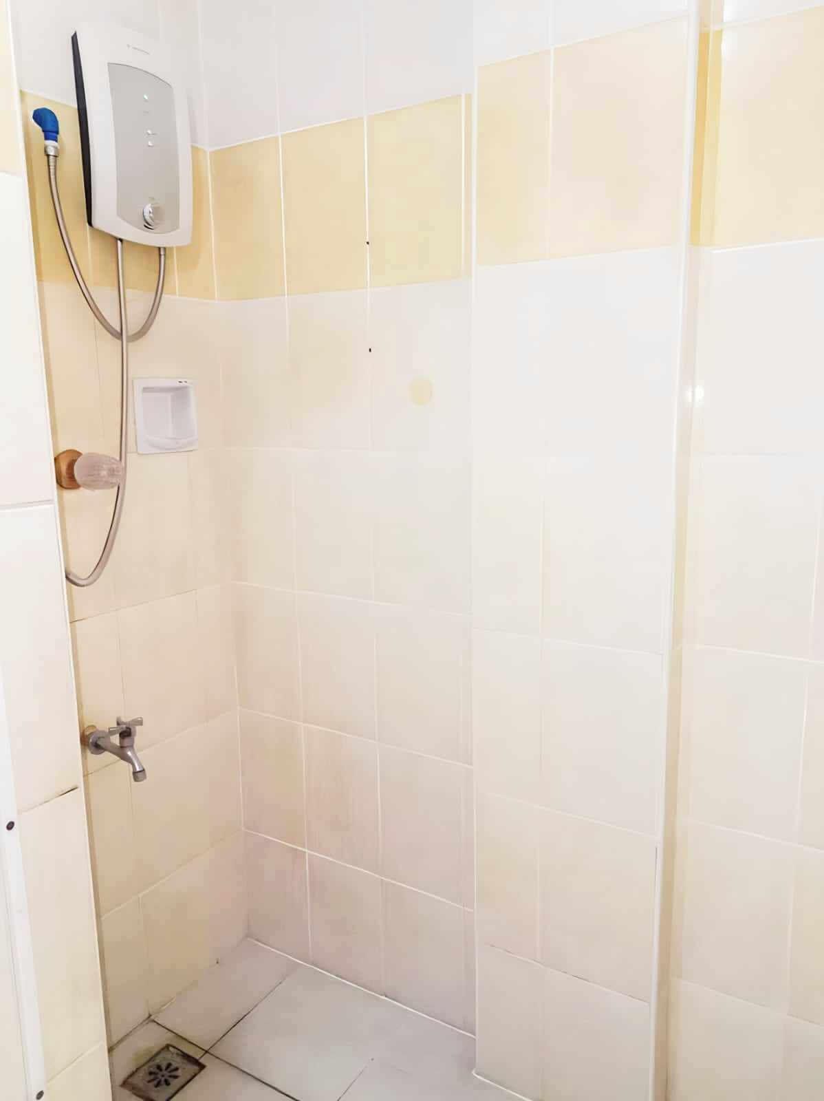 Un petit espace de douche carrelé, équipé d’un pommeau de douche à main, d’un panneau de commande fixé au mur et d’un seul robinet.
